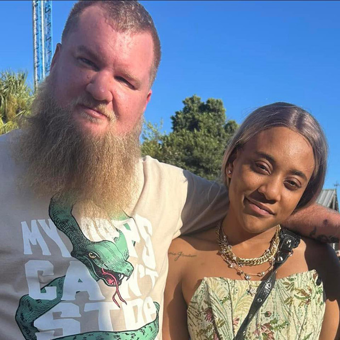 A man and woman standing together outdoors, with trees in the background, in a casual setting.