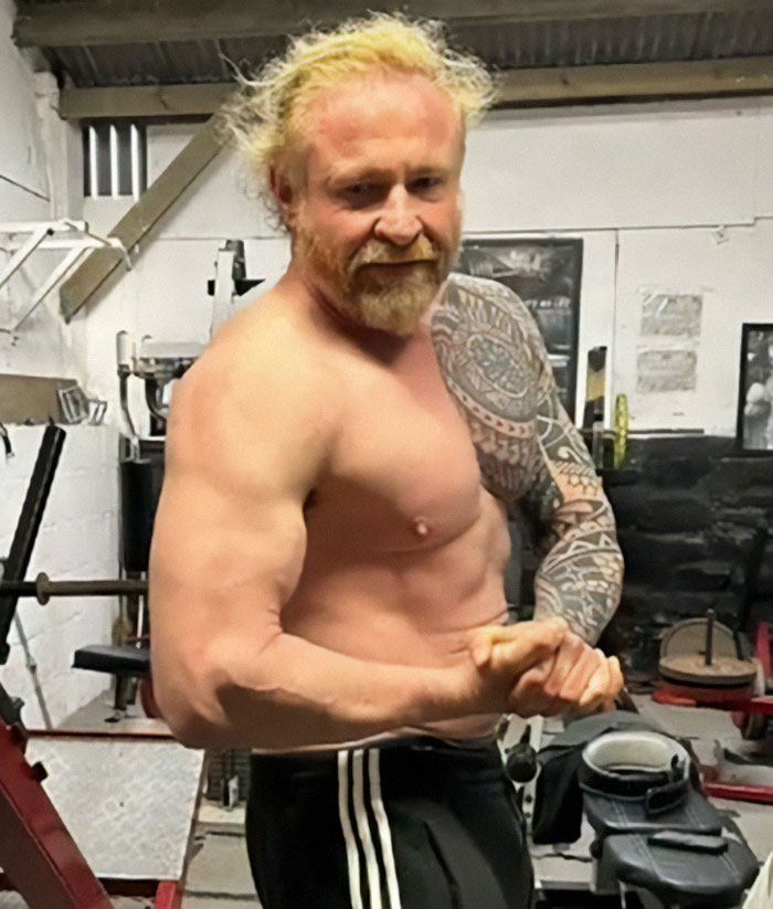 Man flexing muscles in a gym, wearing black pants, representing family tragedy keywords.