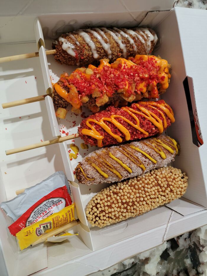A variety of American corn dogs in a box, topped with sauces and crumbs.