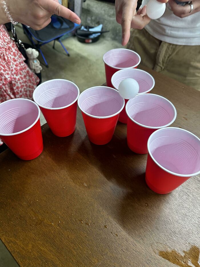 Red cups arranged on a table for a game, with hands holding a ping pong ball; American food culture element.