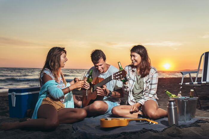 Three friends enjoying a beach sunset, playing guitar, and drinking beer; iconic American tourist vibe.