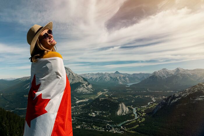 Smiling woman wrapped in Canadian flag, overlooking scenic mountain landscape, representing Americans who moved abroad.