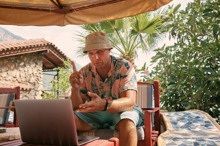 Man in a tropical shirt and bucket hat explaining something while using a laptop outdoors under an umbrella, discussing guys slept married woman why.