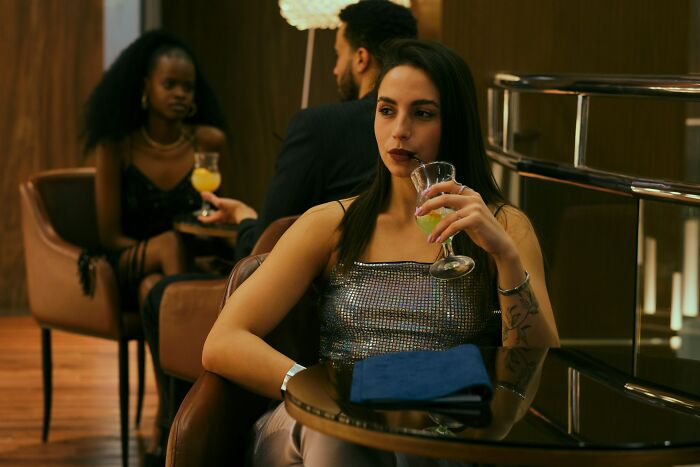 Woman in a sequined top holding a cocktail at a bar, with a couple chatting in the background.