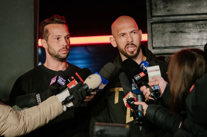 Two men including Andrew Tate speaking to a crowd of reporters, surrounded by microphones and cameras.