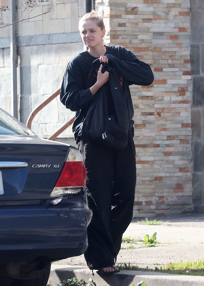 Young person in casual clothing holding a bag, standing next to a parked car on a sunny day.