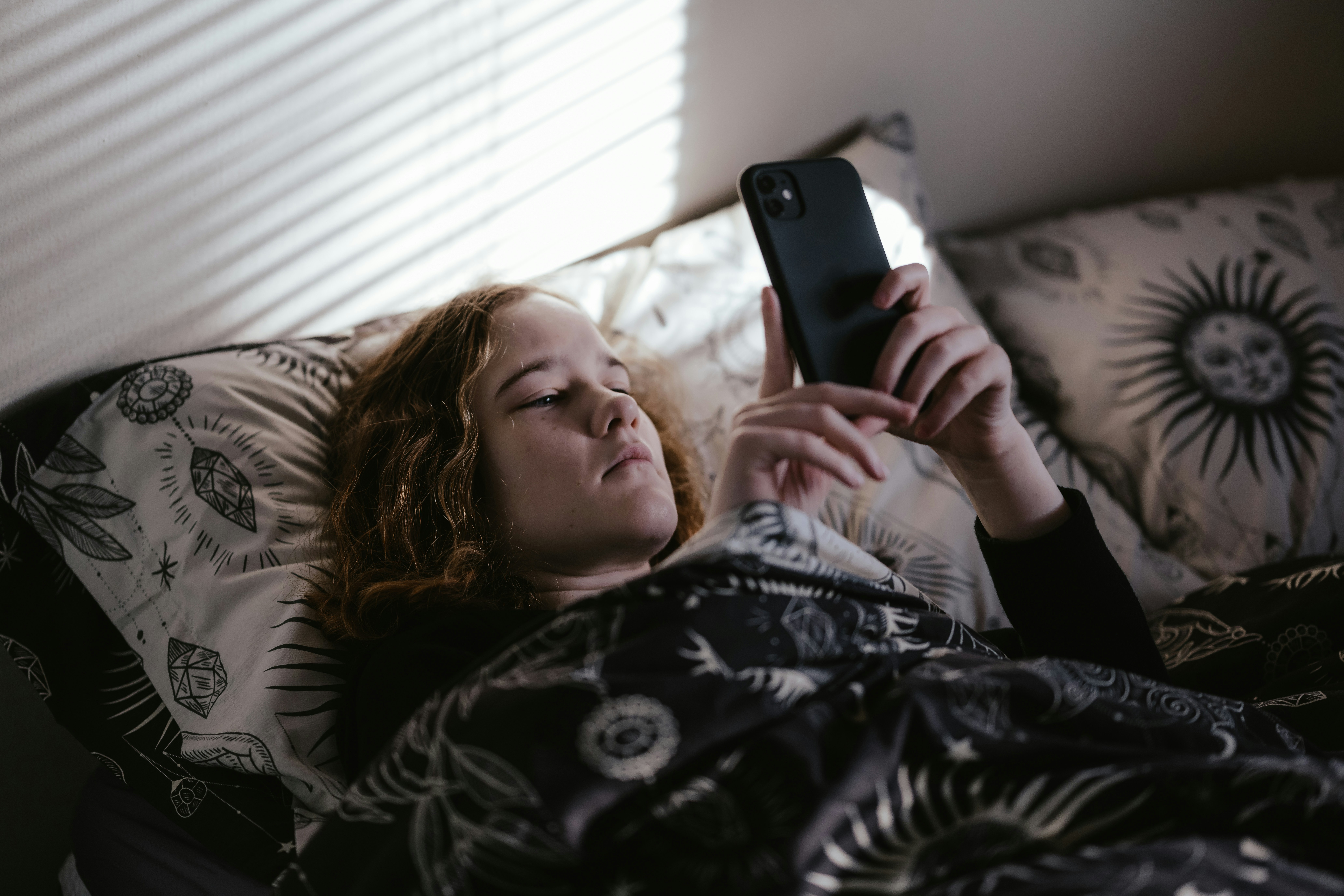 Person lying in bed, focused on a smartphone, under a patterned blanket with sun and moon designs.