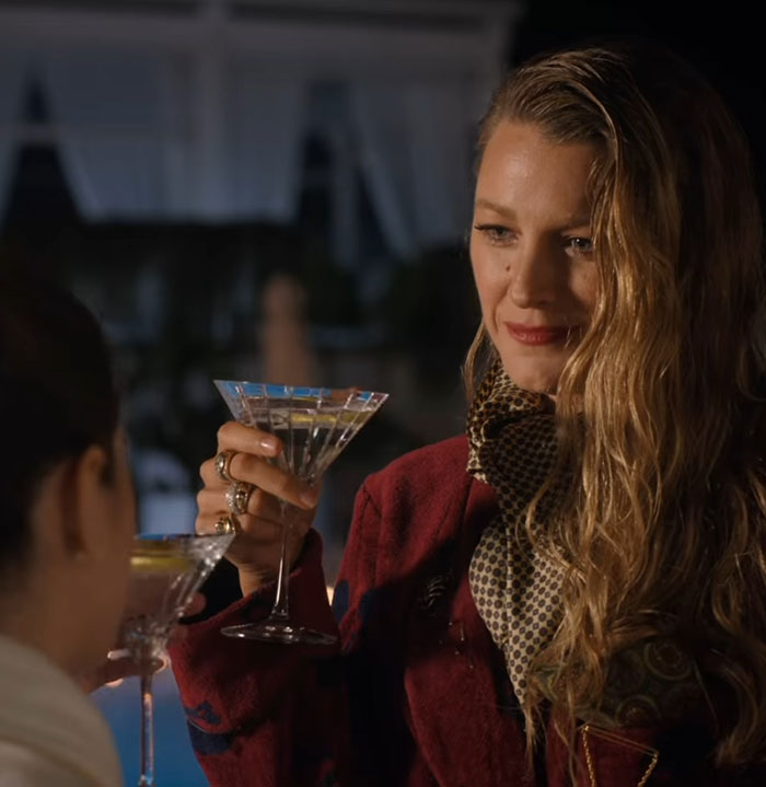 Blonde woman holding a cocktail, wearing a red coat, amid talk about spray tan mishap at a stylish evening event.