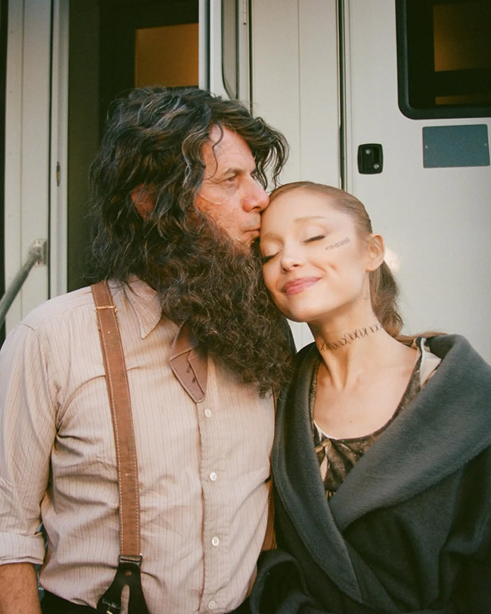 Ariana Grande showcases tattoos and new hair, smiling with a man in costume outside.