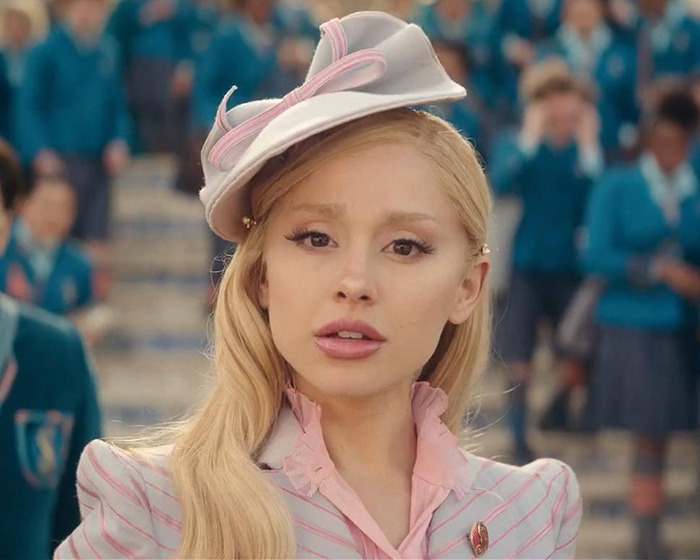 Ariana Grande with new hairstyle and tattoos, standing in a crowd, wearing a stylish hat.