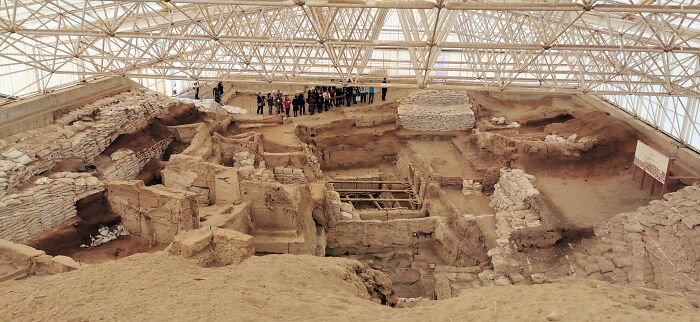 Ancient man-made structures excavation site under protective canopy with tourists observing historical ruins up close.
