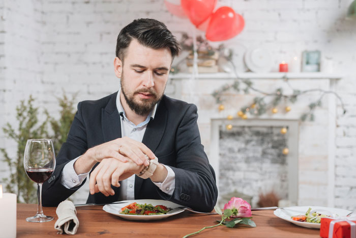 Man dressed in suit checking watch while sitting alone at dinner table with wine glass and rose nearby.