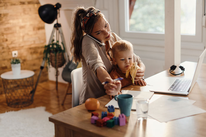 Woman babysitting sister&rsquo;s kids, working on laptop at dining table while toddler eats a banana and plays with toys.