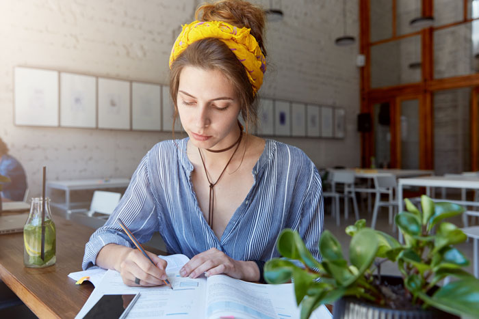 Young woman babysitting sister&rsquo;s kids while working on paperwork at a table with plants and a drink nearby.