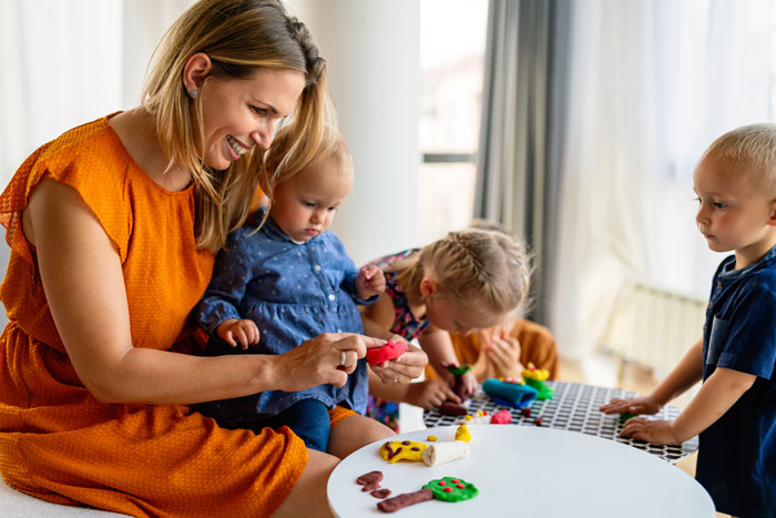 Woman babysitting newborn and children indoors, engaging them with toys and play activities at a table.