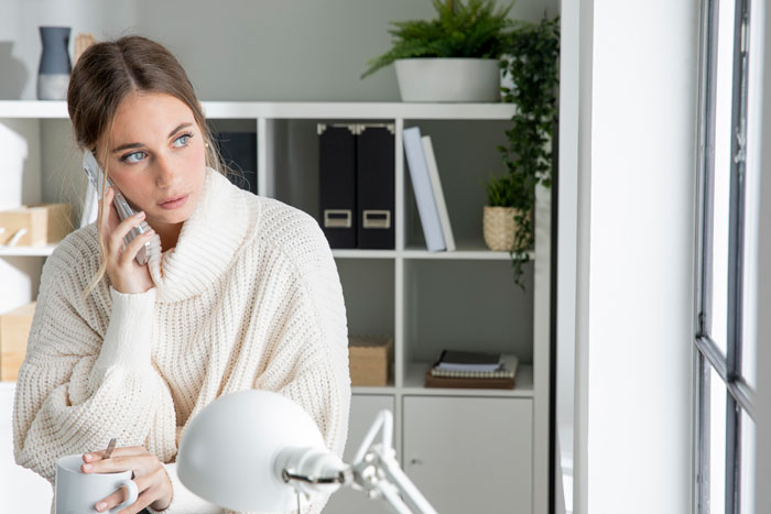 Woman in white sweater looking concerned while talking on phone, holding a mug, near a window in a cozy room.