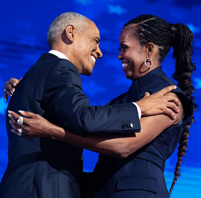Former president, smiling, embraces his wife on stage during a public appearance.