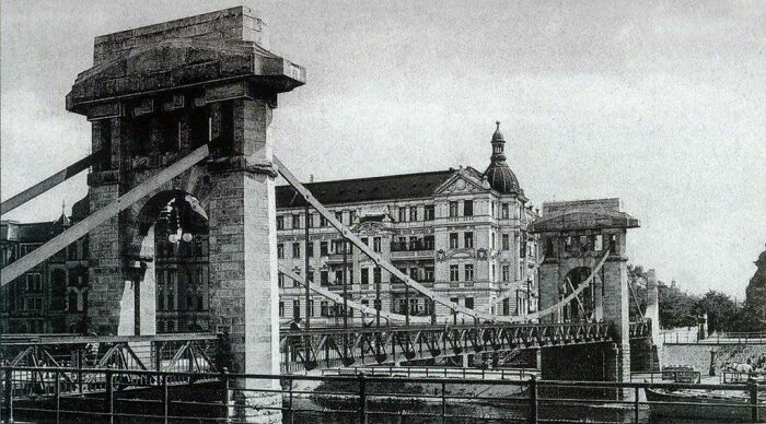 Historic black and white photo of a European suspension bridge with old buildings, showing life in Europe 100 years ago.