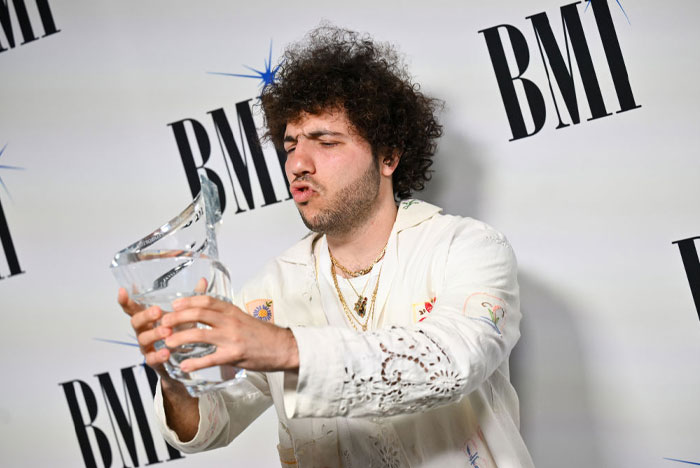 Benny Blanco holding award at BMI event, wearing a white shirt, showcasing successful career and wealth.