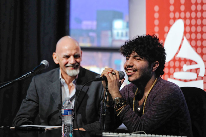 Benny Blanco speaking at an event, holding a microphone, with a man beside him, featuring Grammy signage in the background.