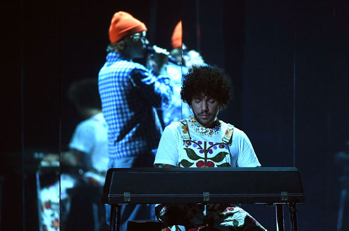 Benny Blanco at a keyboard on stage with blurred singer in background, capturing Benny Blanco's hitmaker vibe.
