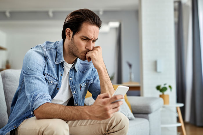 Man wearing denim shirt sitting on couch looking stressed while holding smartphone, illustrating sleep deprivation effects. Man wearing denim shirt sitting on couch looking stressed while holding smartphone, illustrating sleep deprivation effects.