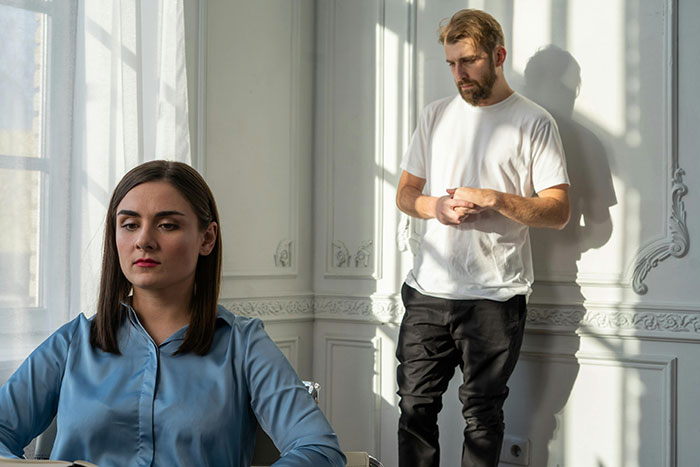 Woman in a blue shirt feeling trapped at home with her boyfriend challenging her medical expertise, in a sunlit room.