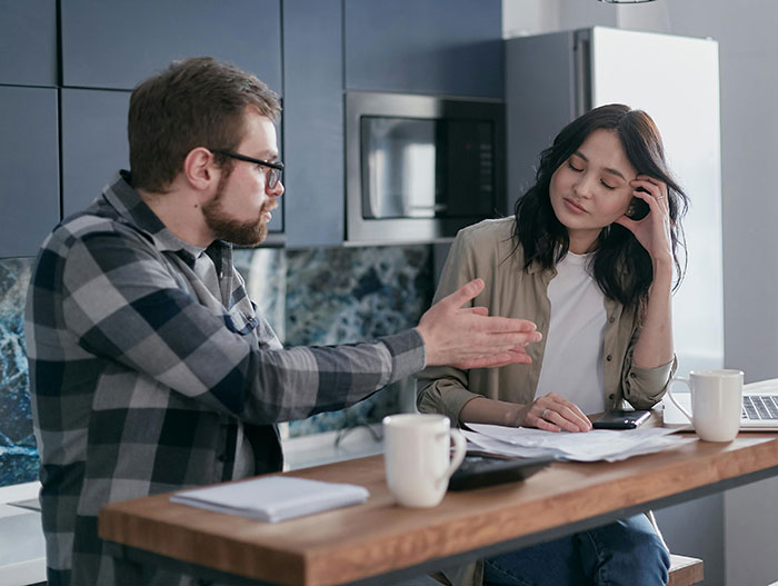Man discussing at a kitchen table, woman with hand on face, appearing frustrated about medical expertise challenge.