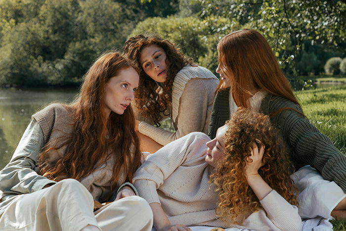 Four women with curly hair conversing on a sunny day by a lake, upset emotions visible in the group dynamic. Four women with curly hair conversing on a sunny day by a lake, upset emotions visible in the group dynamic.