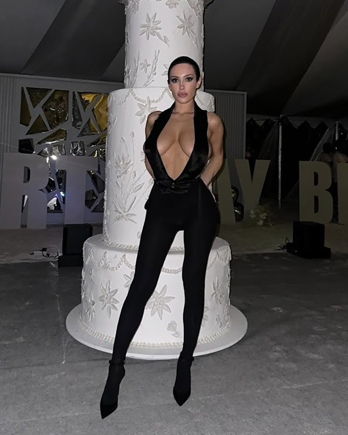 Person in a revealing black outfit standing in front of a large ornate cake.