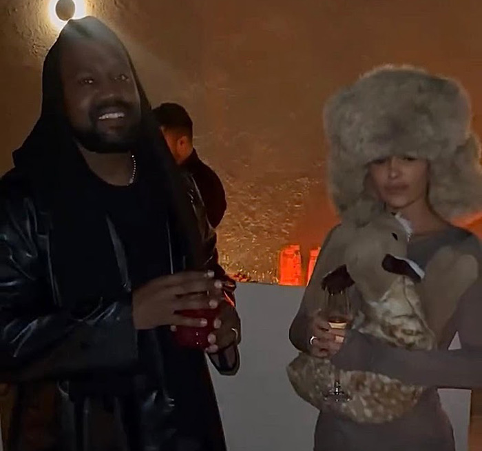 Party scene with Bianca Censori in a furry hat holding a drink, next to a man in a dark outfit, in a dimly lit setting.