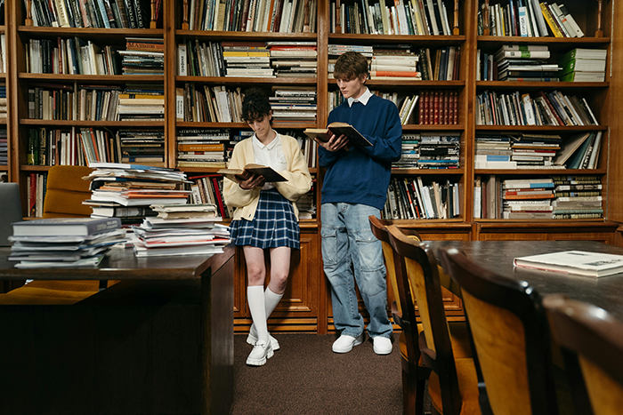 Two students reading in a library, surrounded by bookshelves and stacks of books.