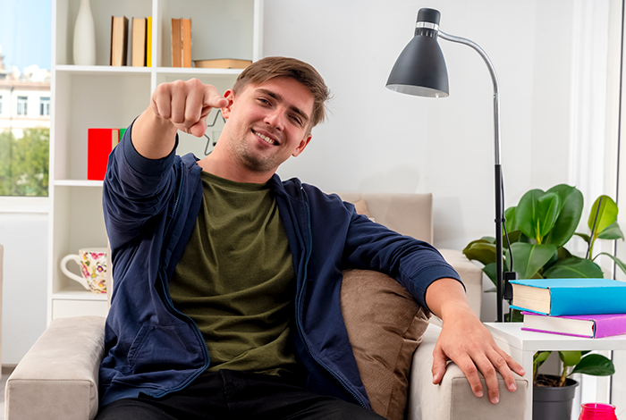 Young man in a cozy room points playfully at the camera, sitting on a couch near a lamp and bookshelf.
