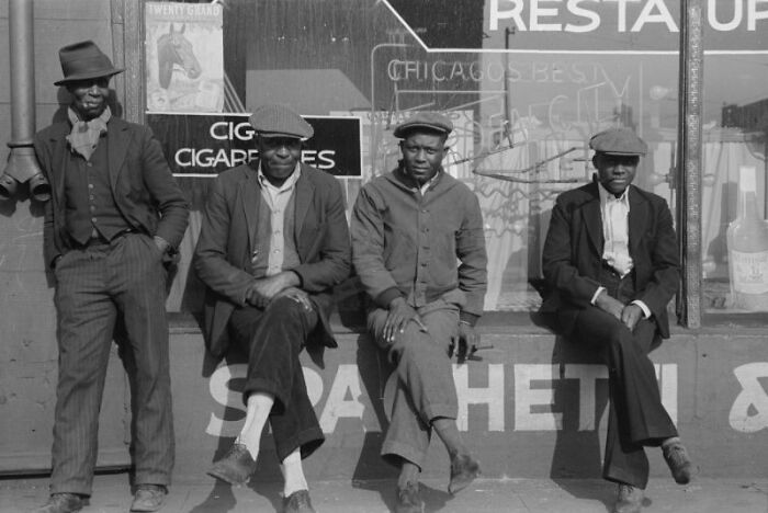 Four African American men dressed in early 20th century clothing sitting and standing outside a storefront.