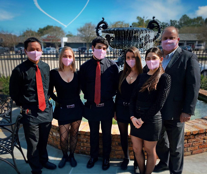 Restaurant staff in matching black outfits and pink masks pose by a fountain under a clear sky.