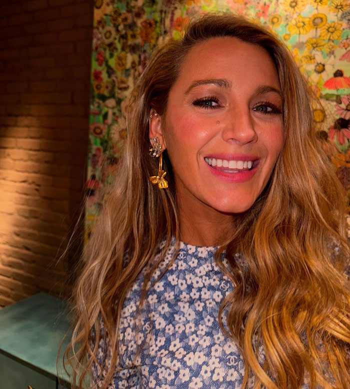 Woman smiling with floral earrings, wearing a blue patterned dress against a colorful floral backdrop.