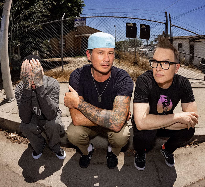 Blink-182's Mark Hoppus sits on curb with bandmates in casual attire, outdoors near a fence.