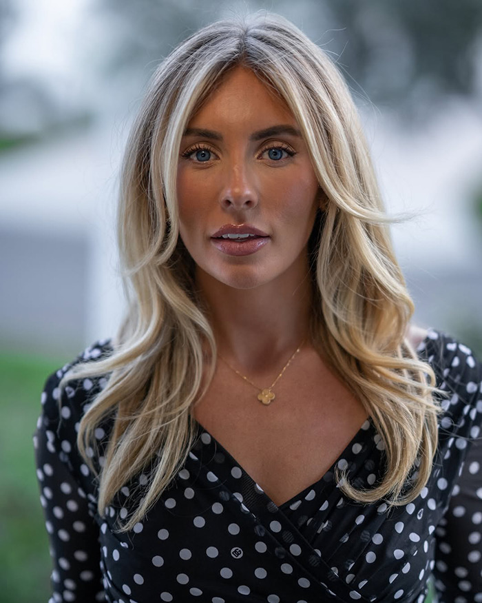 Blonde woman with blue eyes wearing a black polka dot dress, representing Bonnie Blue escorted from soccer stadium.