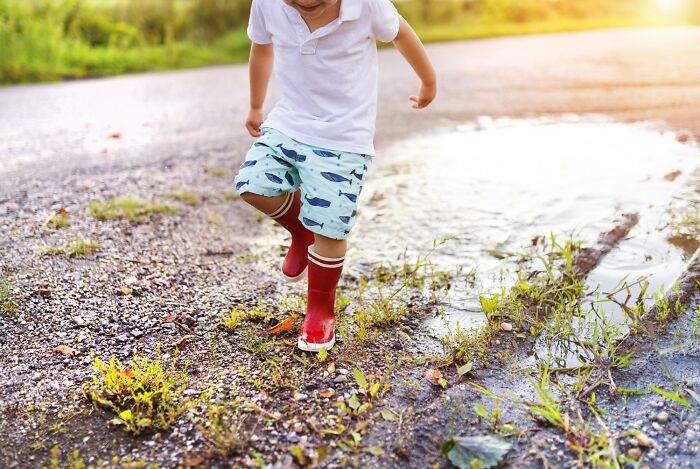 Child in red boots joyfully splashes in a puddle, embodying comedic innocence outdoors.