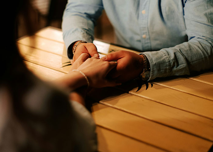 A couple holding hands across a table in a dimly lit setting, suggesting a reconciliation or deep conversation.