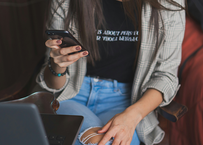 A woman in a plaid jacket, using her phone while sitting in front of a laptop, representing relationship challenges.