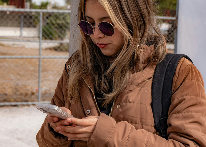 Woman wearing sunglasses using her phone outdoors, bundled in a brown jacket.