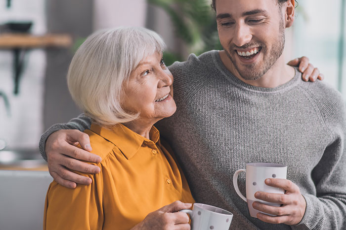Older woman and younger man smiling and holding mugs, illustrating a mom jokingly stealing girlfriend's seat to separate couple.