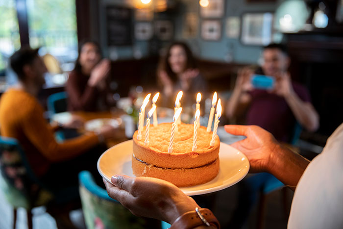 Birthday cake with lit candles being served to a group of friends, illustrating couple breaks up after mom jokingly steals GF&rsquo;s seat.