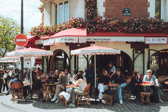 People enjoying a sunny day at a European cafe with outdoor seating under umbrellas.
