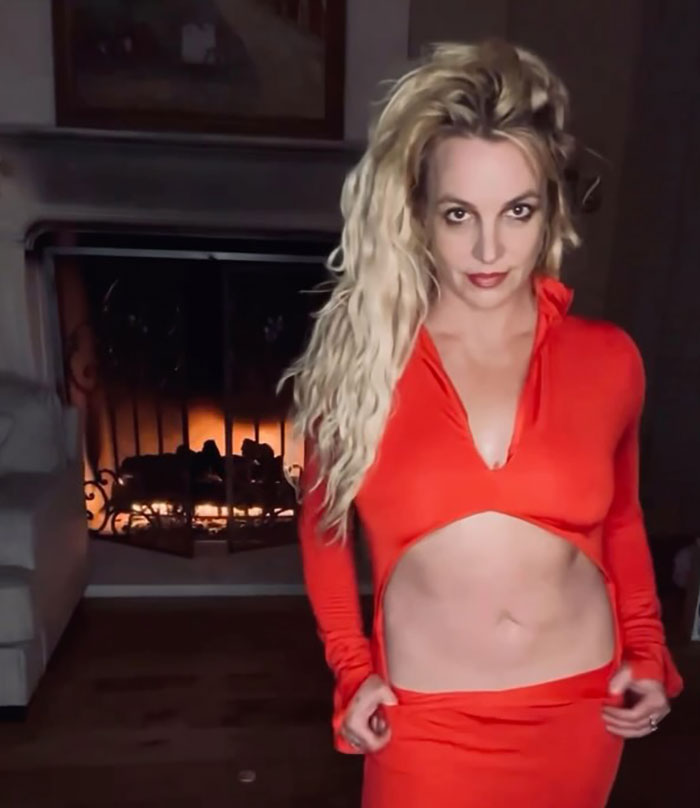 Blonde woman in a red outfit standing confidently near a fireplace in a dimly lit room.