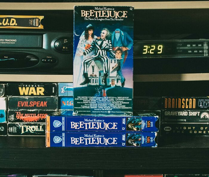 VHS tapes including "Beetlejuice" displayed on a shelf beside a retro clock and video player, featuring loopholes nostalgia.