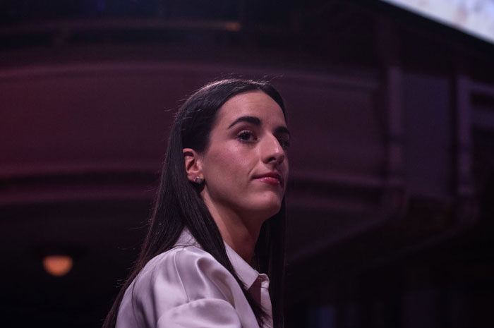 Caitlin Clark in a white shirt under moody lighting, representing rising net worth and sponsorship interest.
