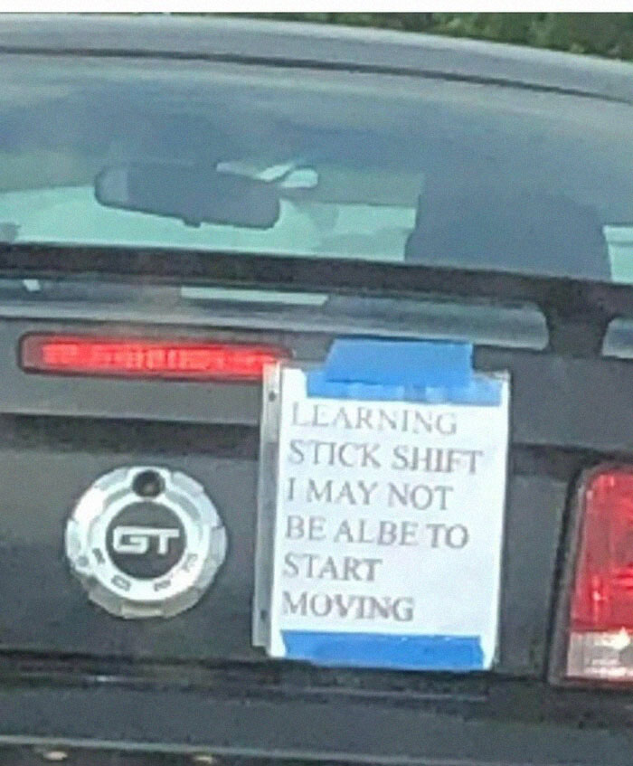 Car meme showing a sign on a vehicle: "Learning stick shift, may not start moving."