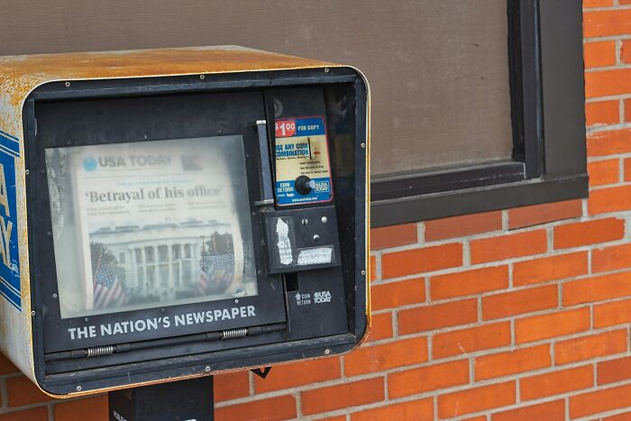 Newspaper vending box with headline about an office betrayal, next to a brick wall, showcasing exploited loopholes.